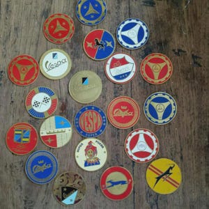May include: A collection of twenty-two circular metal emblems and badges featuring various colorful designs, including the Vespa logo, a jester illustration, a leaping gazelle, checkered flags, and geometric patterns in red, blue, gold, and white.