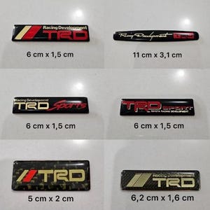 Op de afbeelding: Zes rechthoekige auto-emblemen met TRD Racing Development branding in zwart, rood en goud. De afmetingen zijn 6 cm bij 1,5 cm, 11 cm bij 3,1 cm, 6 cm bij 1,5 cm, 6 cm bij 1,5 cm, 5 cm bij 2 cm en 6,2 cm bij 1,6 cm.