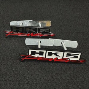 Könnte beinhalten: Zwei HKS Power Embleme mit einem rechteckigen schwarz-weißen Logo und rotem, stilisiertem Text darunter. Jedes Emblem hat zwei Metallstifte auf der Rückseite zur Befestigung. Das Design ist markant und zeigt den Markennamen HKS deutlich.