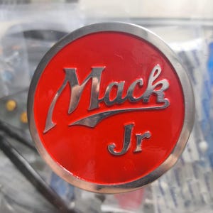 May include: A round, red emblem with the words "Mack Jr" in silver lettering. The emblem has a silver border and is likely a logo or badge.