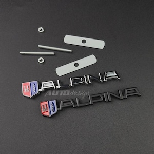 May include: Two Alpina car emblems, one in chrome and one in matte black, each featuring a shield logo with red, blue, and white details. The set includes two threaded metal rods, two small hexagonal nuts, and two rectangular metal mounting plates.