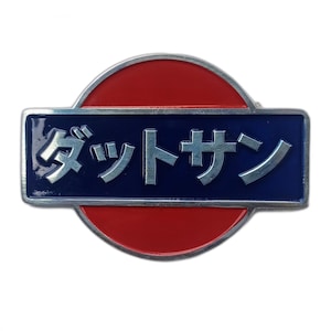May include: A Datsun emblem pin with Japanese text. The pin features a rectangular blue section with silver Japanese characters, set against a red circular background. The pin has a silver border.