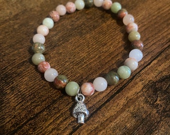 Puede incluir: Un brazalete hecho de cuentas rosas, verdes y marrones con un dije de hongo plateado.