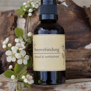 May include: A dark amber glass spray bottle with a black plastic pump top. The beige label features the text "Herzverbindung" and "öffnend & verbindend" in black font, accompanied by a small botanical illustration of a flower on the right side.