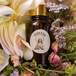 May include: A dark amber glass dropper bottle with a gold collar and white cap. The label features the text GÖTTIN above an illustration of a figure with a halo of stars. The bottle is surrounded by pale yellow flowers and purple yarrow.