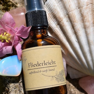 May include: An amber glass spray bottle with a black pump top and a tan paper label. The label features the text Fliederleicht, aufhellend & sanft lösend, and a botanical illustration of a lilac flower cluster. The bottle is surrounded by a pink flower.