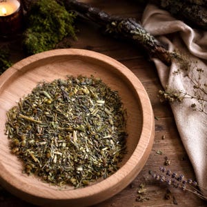 May include: A round, light-colored wooden bowl filled with a loose-leaf herbal tea blend. The mixture contains various dried green herbs, small yellow flower buds, and purple lavender sprigs, creating a textured, earthy, and botanical appearance.