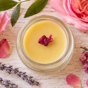 May include: A small, clear glass jar filled with a smooth, pale yellow balm. A single dried rose petal rests on the surface of the balm. The jar is surrounded by fresh pink roses, sprigs of purple lavender, and small clusters of pink flowers.