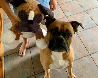 Puede incluir: Un juguete de perro tejido en beige y blanco, sostenido por una persona, junto a un perro Boxer real. El juguete tiene orejas y hocico de color marrón oscuro. El perro real es beige con pecho blanco y hocico negro.