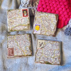 May include: Four square ceramic coasters with a vintage map design. Each coaster features a different vintage postage stamp with images of a rose, a man, a woman, and a bird. The coasters are on a gray fuzzy surface.