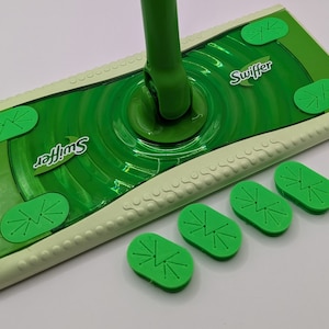 Puede incluir: Una mopa Swiffer verde con un mango verde. El cabezal de la mopa es rectangular con un borde verde claro y la palabra "Swiffer" impresa. Se incluyen cuatro almohadillas verdes.