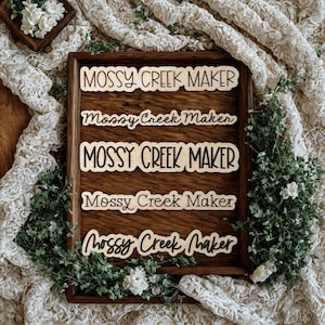 以下が含まれることがあります： 木製のトレイには、さまざまなフォントで「MOSSY CREEK MAKER」というテキストが書かれた5つの木製切り抜きが表示されています。 トレイは緑と白い花に囲まれ、クリーム色のテクスチャードファブリックを背景にしています。