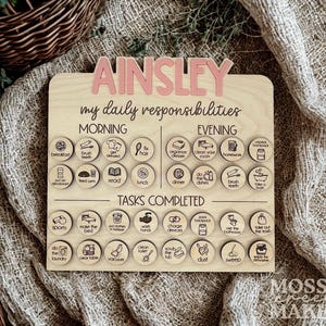 May include: Wooden daily responsibility chart with the name "AINSLEY" in pink letters. The chart includes morning and evening tasks, with icons for each activity. The words "my daily responsibilities" are also visible.