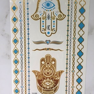 May include: Gold and blue temporary tattoo sheet with two hamsa hand designs, each with an eye in the center. The sheet also includes several decorative borders with chains and geometric shapes.