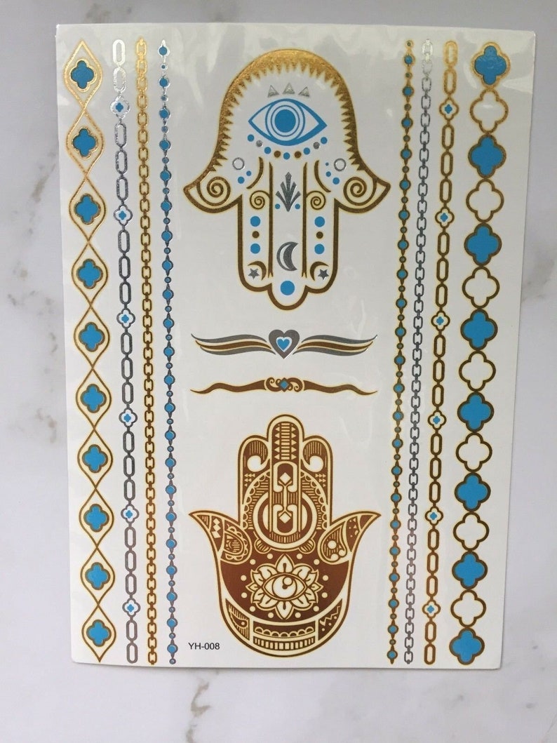 May include: Gold and blue temporary tattoo sheet with two hamsa hand designs, each with an eye in the center. The sheet also includes several decorative borders with chains and geometric shapes.