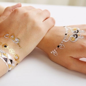 May include: Two hands with gold and silver temporary tattoos. The tattoos are intricate designs with swirls and hearts.