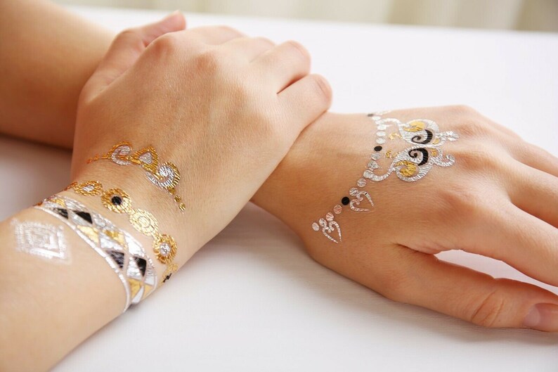 May include: Two hands with gold and silver temporary tattoos. The tattoos are intricate designs with swirls and hearts.