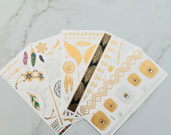 May include: A set of six gold and white temporary tattoos featuring various designs, including feathers, dreamcatchers, wings, and geometric patterns. The tattoos are on white sheets with a white background.