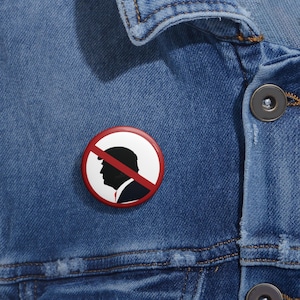 Puede incluir: Un botón rojo y blanco con una silueta de una cabeza de persona con una línea roja que la atraviesa. El texto "No" está escrito en blanco en el botón.