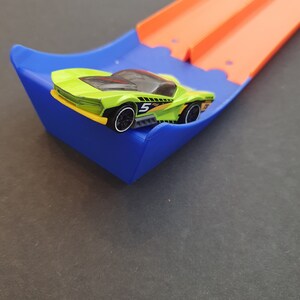 May include: A green toy car with a black number 5 on the side is on a blue and orange toy race track.