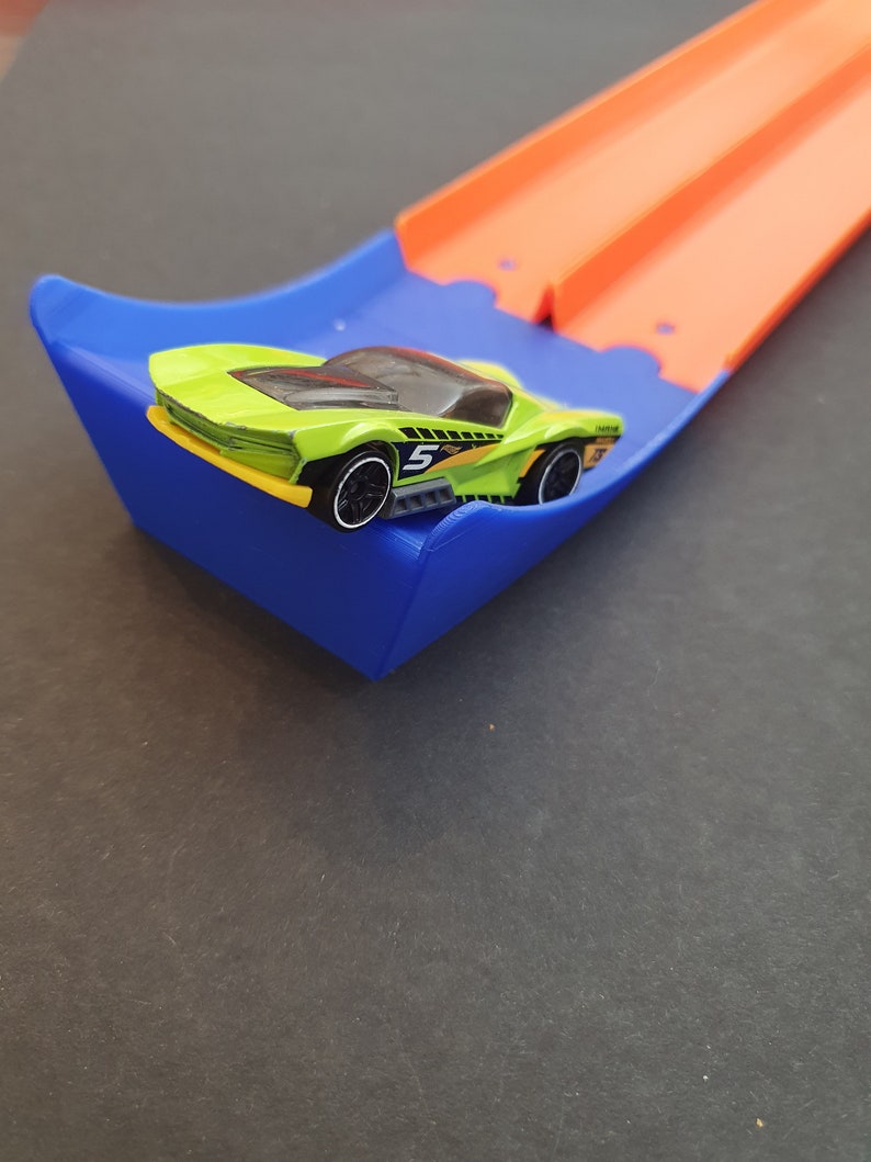 May include: A green toy car with a black number 5 on the side is on a blue and orange toy race track.