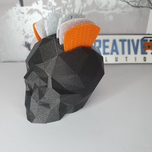 Puede incluir: Un macetero negro con forma de calavera geom&eacute;trica con una superficie texturizada. El macetero est&aacute; lleno de peque&ntilde;os objetos decorativos blancos y naranjas.
