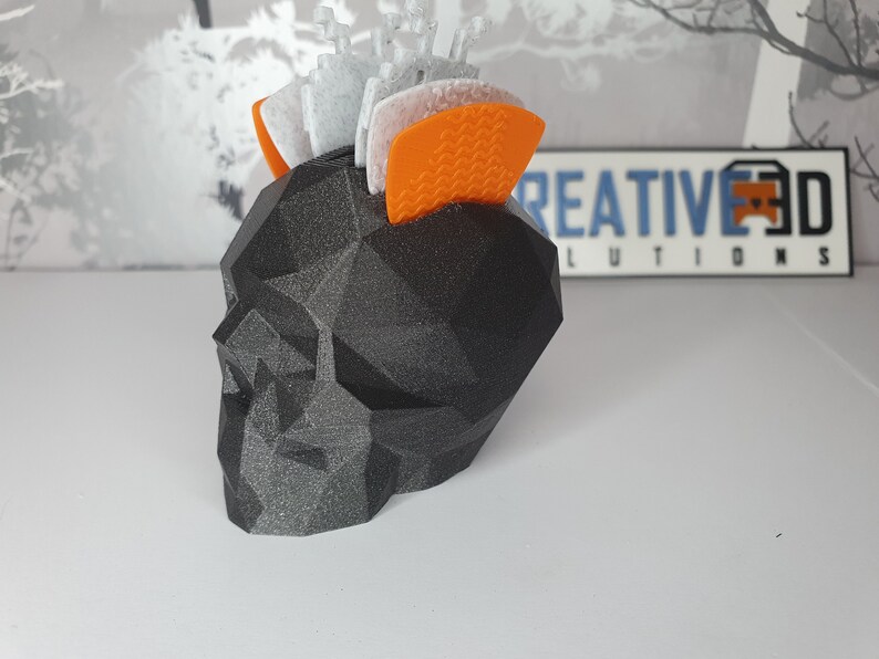 Puede incluir: Un macetero negro con forma de calavera geom&eacute;trica con una superficie texturizada. El macetero est&aacute; lleno de peque&ntilde;os objetos decorativos blancos y naranjas.