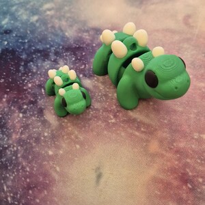 May include: Two green dinosaur figurines with white spots. The larger dinosaur is standing on its hind legs, while the smaller dinosaur is lying on its belly.