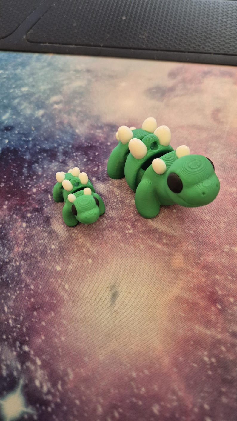 May include: Two green dinosaur figurines with white spots. The larger dinosaur is standing on its hind legs, while the smaller dinosaur is lying on its belly.