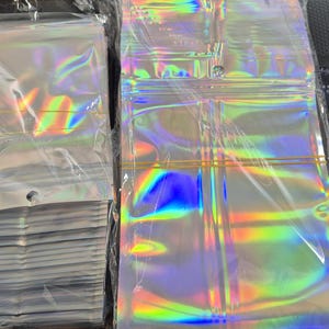 May include: A collection of clear, iridescent zip-top bags. The bags have a holographic finish, displaying a spectrum of colours. Some are stacked, others are flat. Each bag features a zip closure and a small hole at the top for hanging or display.