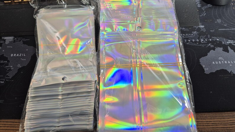 May include: A collection of clear, iridescent zip-top bags. The bags have a holographic finish, displaying a spectrum of colours. Some are stacked, others are flat. Each bag features a zip closure and a small hole at the top for hanging or display.