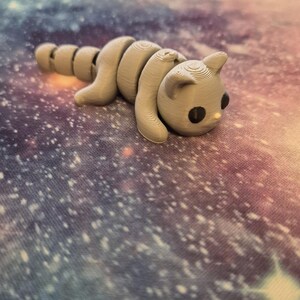 May include: A grey 3D printed toy shaped like a cat with a segmented body. The cat is lying on a colourful surface with a galaxy pattern.