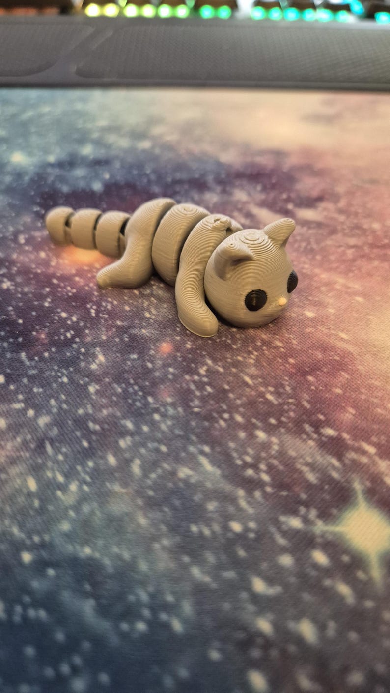 May include: A grey 3D printed toy shaped like a cat with a segmented body. The cat is lying on a colourful surface with a galaxy pattern.