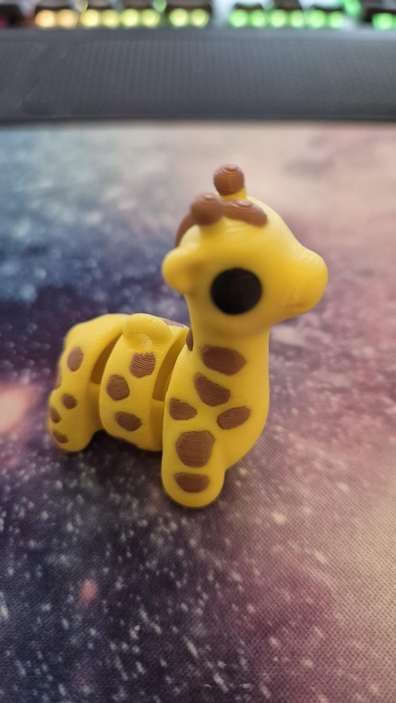 May include: A small yellow and brown giraffe figurine with a segmented body. The giraffe has a large black eye and a brown nose.