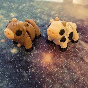 May include: Two 3D-printed cow figurines. One is brown with black accents, the other is white with black spots. Both have small horns and black hooves. They are positioned on a galaxy-themed surface.