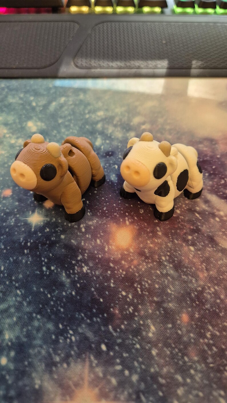 May include: Two 3D-printed cow figurines. One is brown with black accents, the other is white with black spots. Both have small horns and black hooves. They are positioned on a galaxy-themed surface.