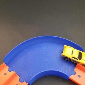 May include: A blue plastic track piece for a toy car race set. The track piece is curved and has a yellow toy car driving on it.