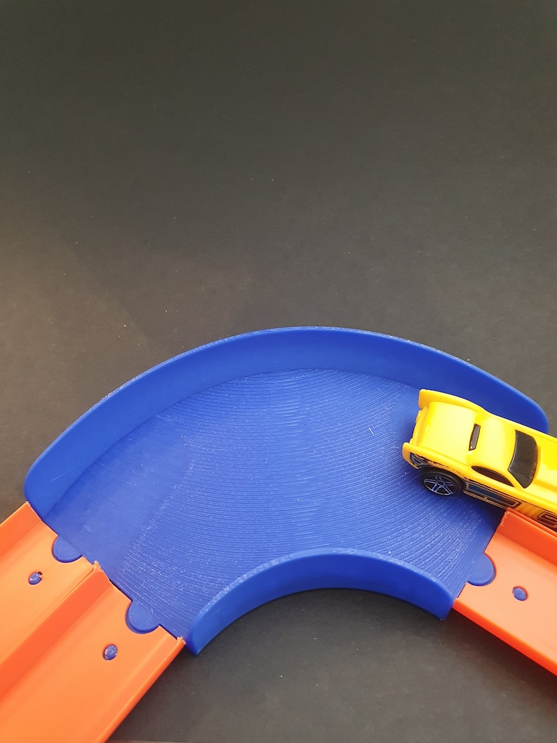 May include: A blue plastic track piece for a toy car race set. The track piece is curved and has a yellow toy car driving on it.