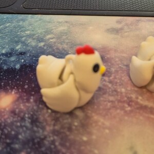 May include: Two white ceramic chicken figurines with red combs and black eyes. The chicken on the left has its wings spread out. The chickens are on a white and purple background.