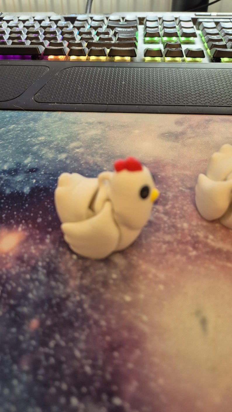 May include: Two white ceramic chicken figurines with red combs and black eyes. The chicken on the left has its wings spread out. The chickens are on a white and purple background.