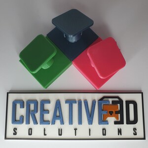 May include: Three 3D printed square blocks with a raised square button on top. The blocks are green, blue, and red. The blocks are arranged in a triangular shape. The text "CREATIVE3D SOLUTIONS" is printed on a white rectangular sign with a black border.