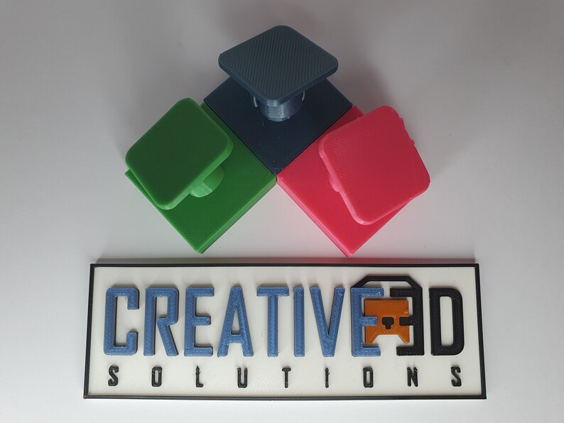 May include: Three 3D printed square blocks with a raised square button on top. The blocks are green, blue, and red. The blocks are arranged in a triangular shape. The text "CREATIVE3D SOLUTIONS" is printed on a white rectangular sign with a black border.