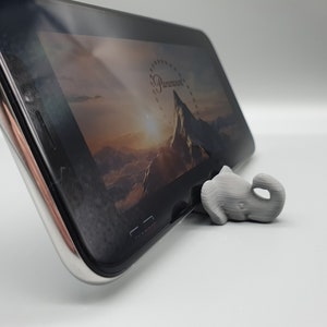 May include: A gray 3D printed elephant figurine is used as a phone stand to prop up a black smartphone. The phone screen shows a mountain range with a sunrise in the background.