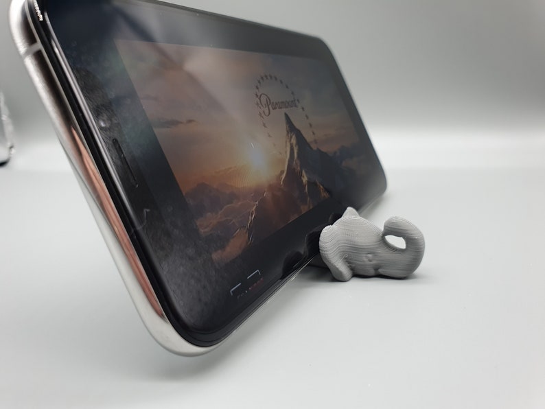 May include: A gray 3D printed elephant figurine is used as a phone stand to prop up a black smartphone. The phone screen shows a mountain range with a sunrise in the background.