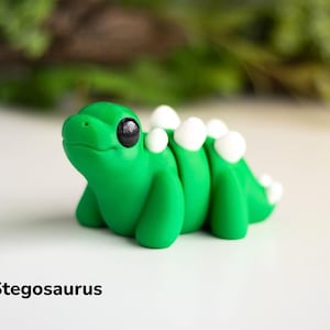 May include: A small, green, hand-crafted baby stegosaurus figurine with white plates on its back. The figurine has black eyes and a cute expression.