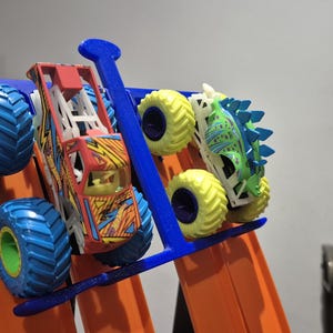 May include: Two toy monster trucks, one red and yellow with blue wheels, the other green and blue with yellow wheels, are displayed on a blue and orange track.