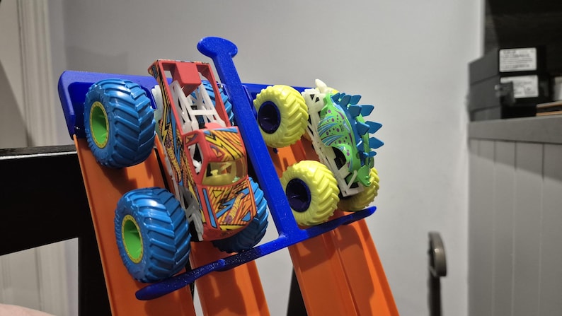 May include: Two toy monster trucks, one red and yellow with blue wheels, the other green and blue with yellow wheels, are displayed on a blue and orange track.