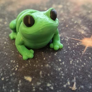 May include: A small green 3D printed frog figurine with black eyes. The frog is sitting on a dark surface with white speckles.