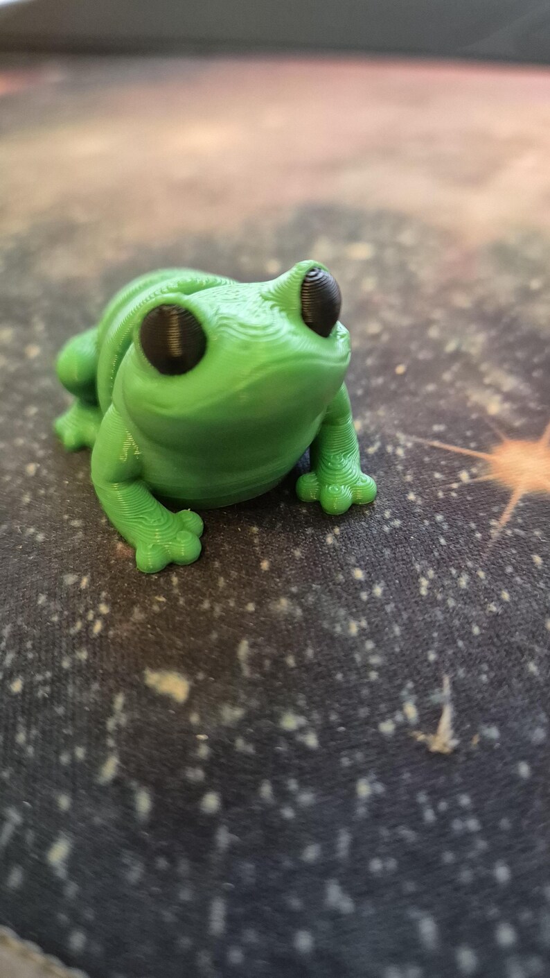 May include: A small green 3D printed frog figurine with black eyes. The frog is sitting on a dark surface with white speckles.