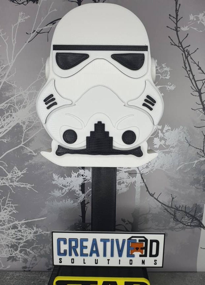 May include: A white 3D printed model of a Star Wars Stormtrooper helmet on a black stand. The stand has a yellow base with the text "CREATIVE 3D SOLUTIONS" on a white plaque.
